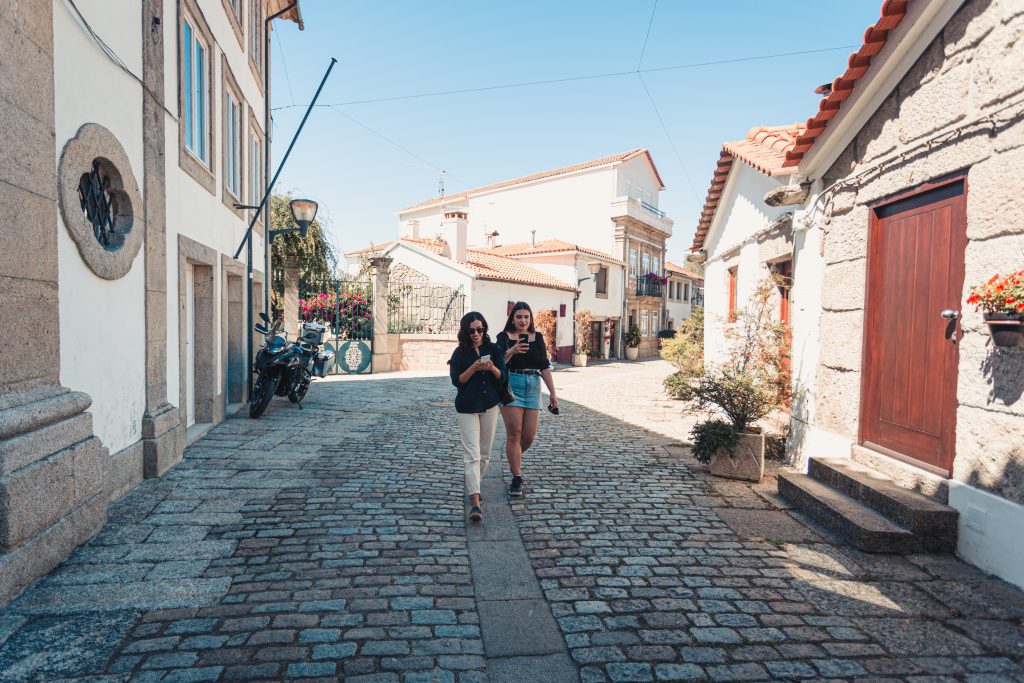 Two people walk down a cobbled street in a sunny, quiet village in Croatia, both looking at their mobiles as they check their travel itinerary.